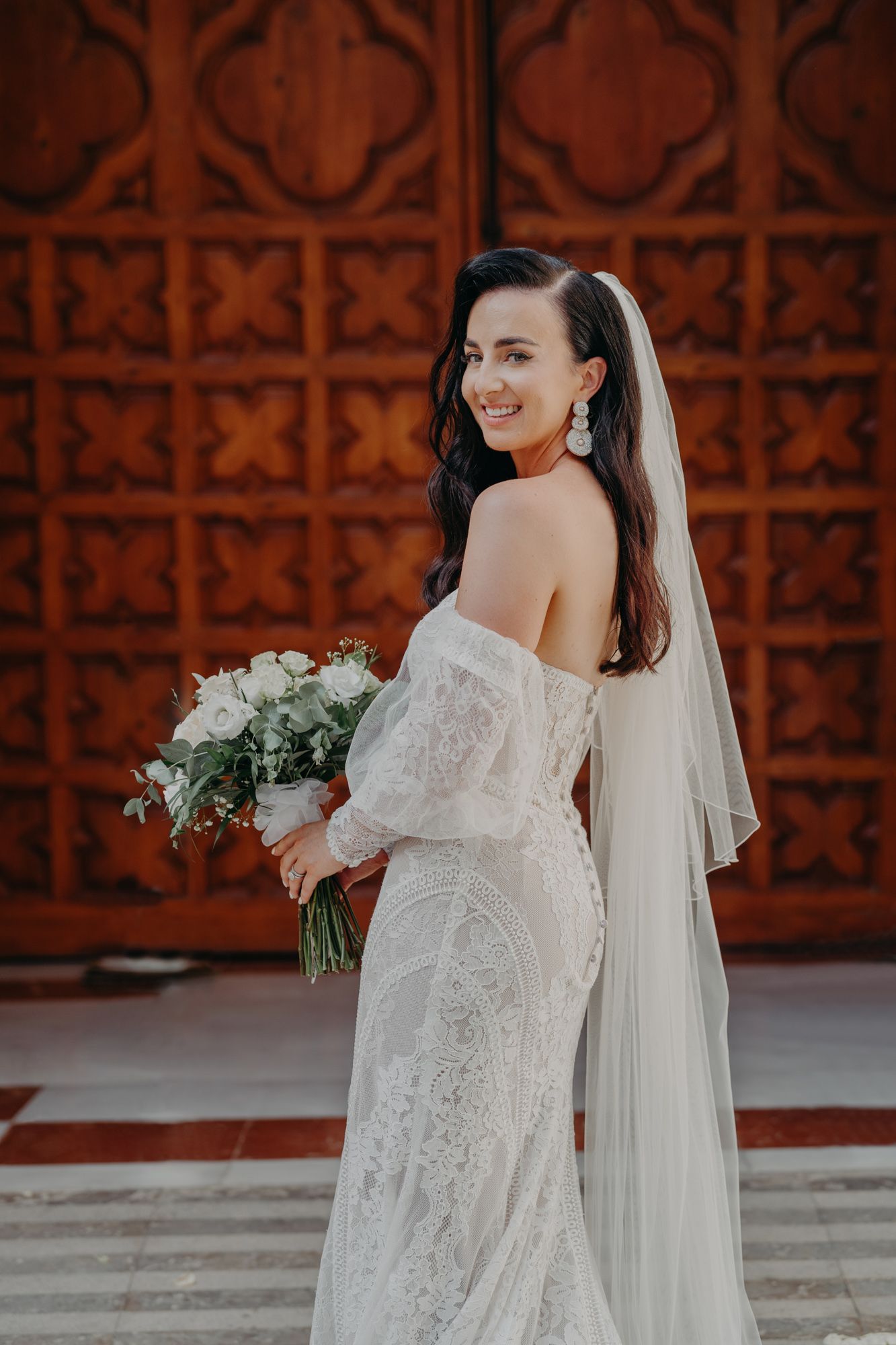 A bride in a lace off-shoulder gown poses with white bouquet against terracotta wall.