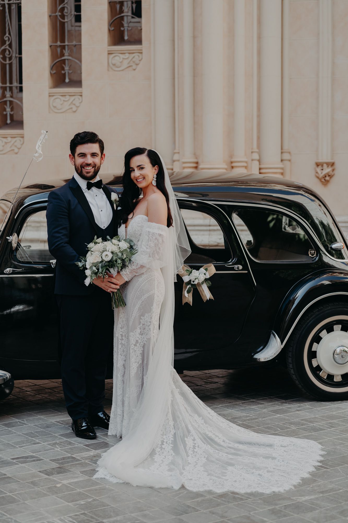 A couple poses with their wedding bouquets beside a classic black vintage car.