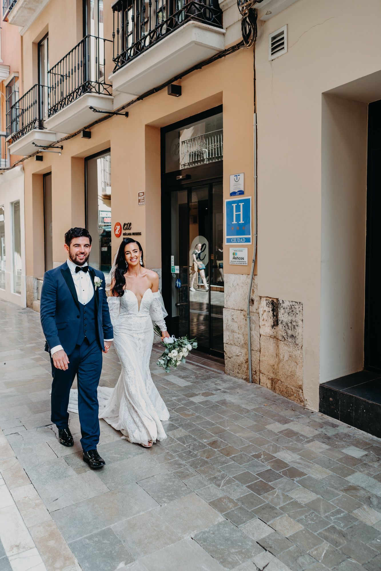 A couple in wedding attire walking outside a hotel entrance on a stone paved street in Europe.