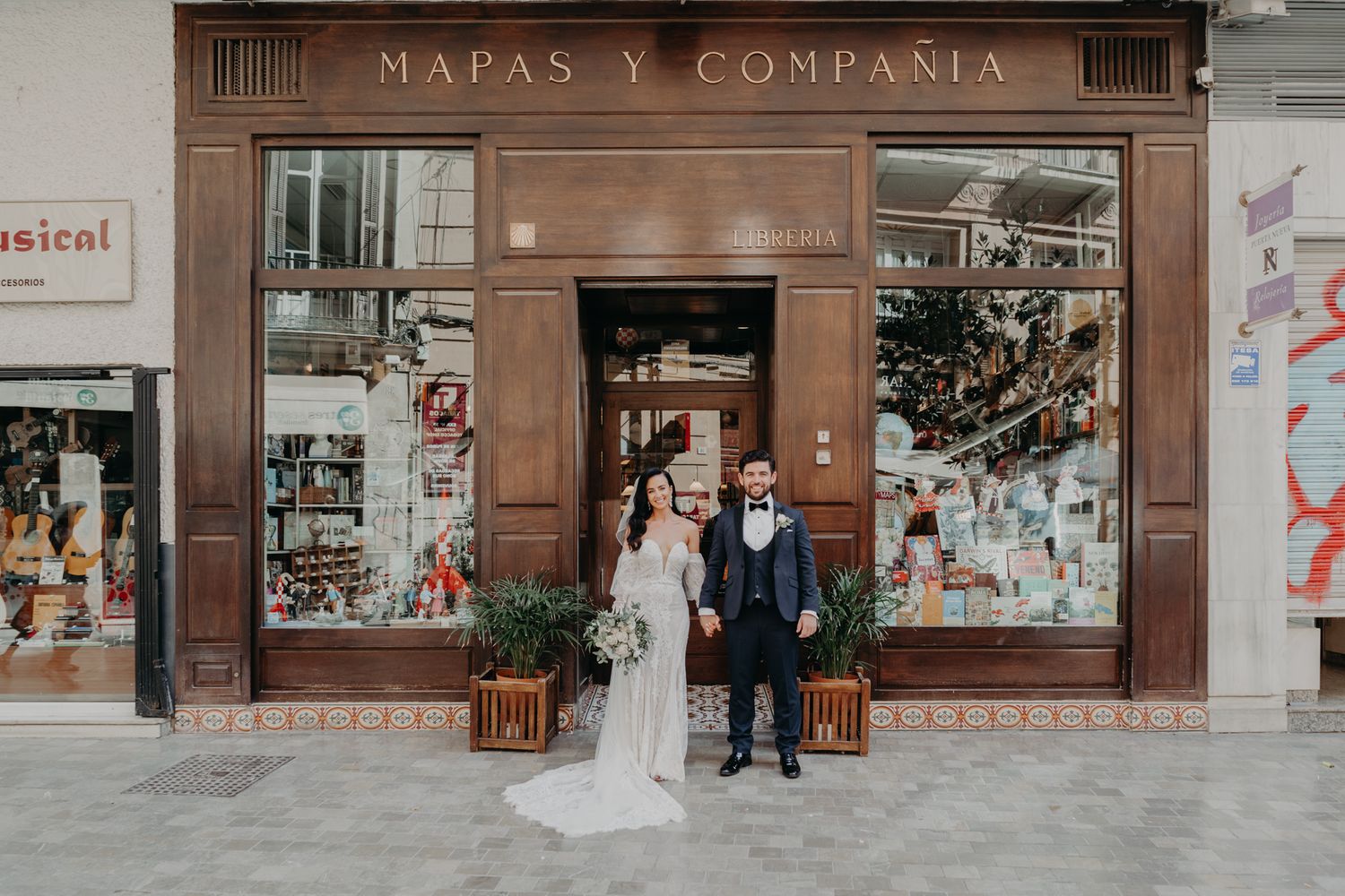 A couple standing in wedding attire in front of a vintage storefront reading 'Mapas y Compania'.