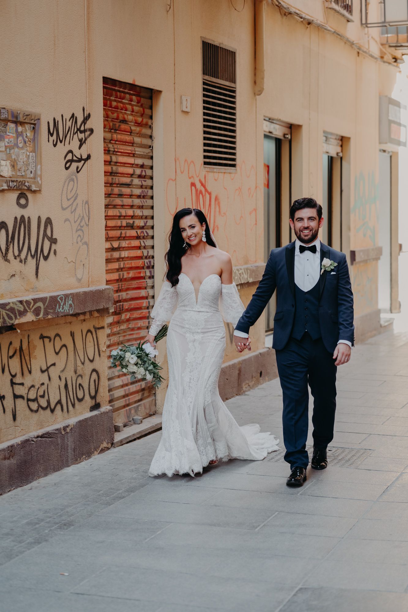 A newlywed couple walks hand in hand down an urban street with graffiti walls behind them.
