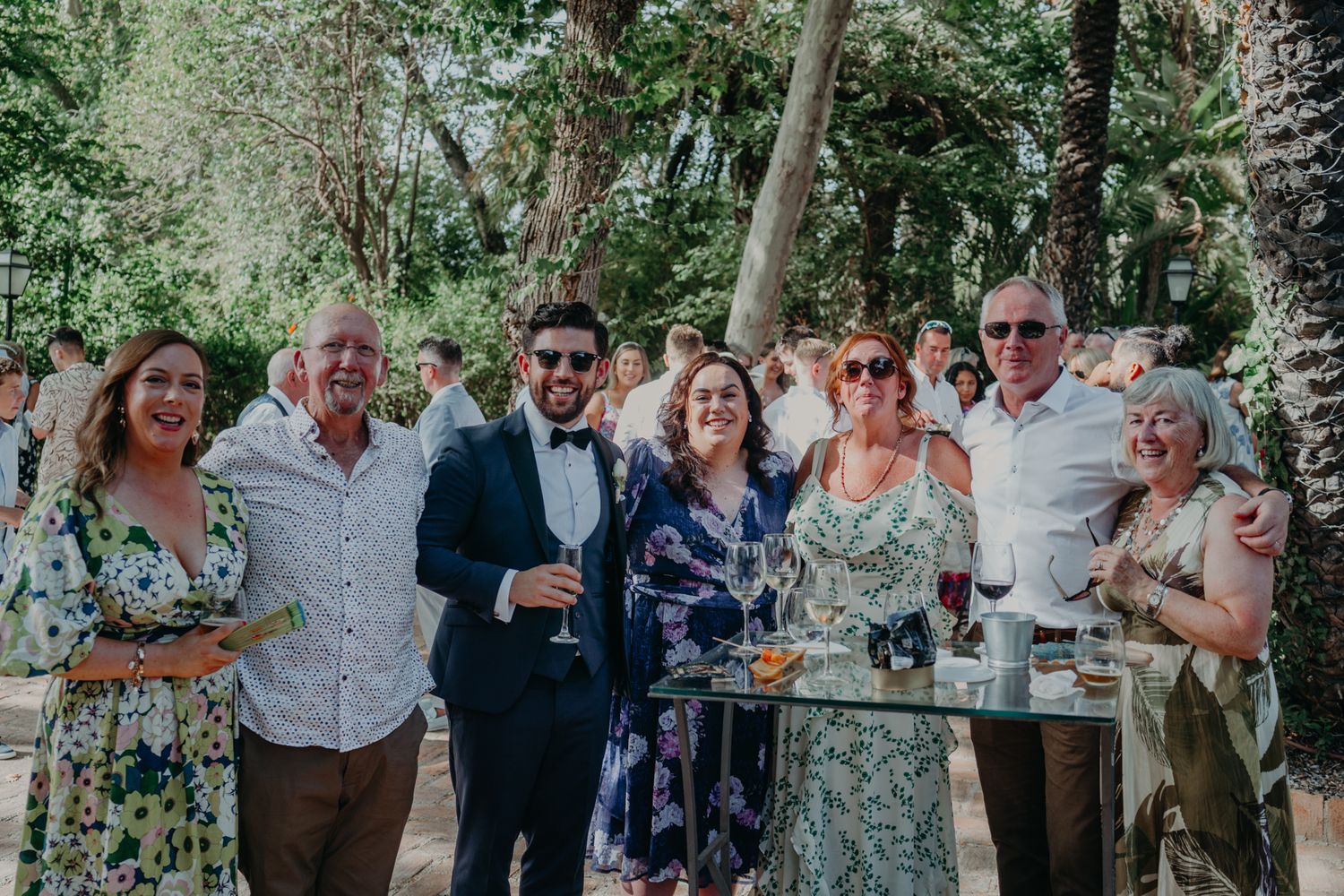 A group stands together at an outdoor garden party holding drinks and smiling on a sunny day.
