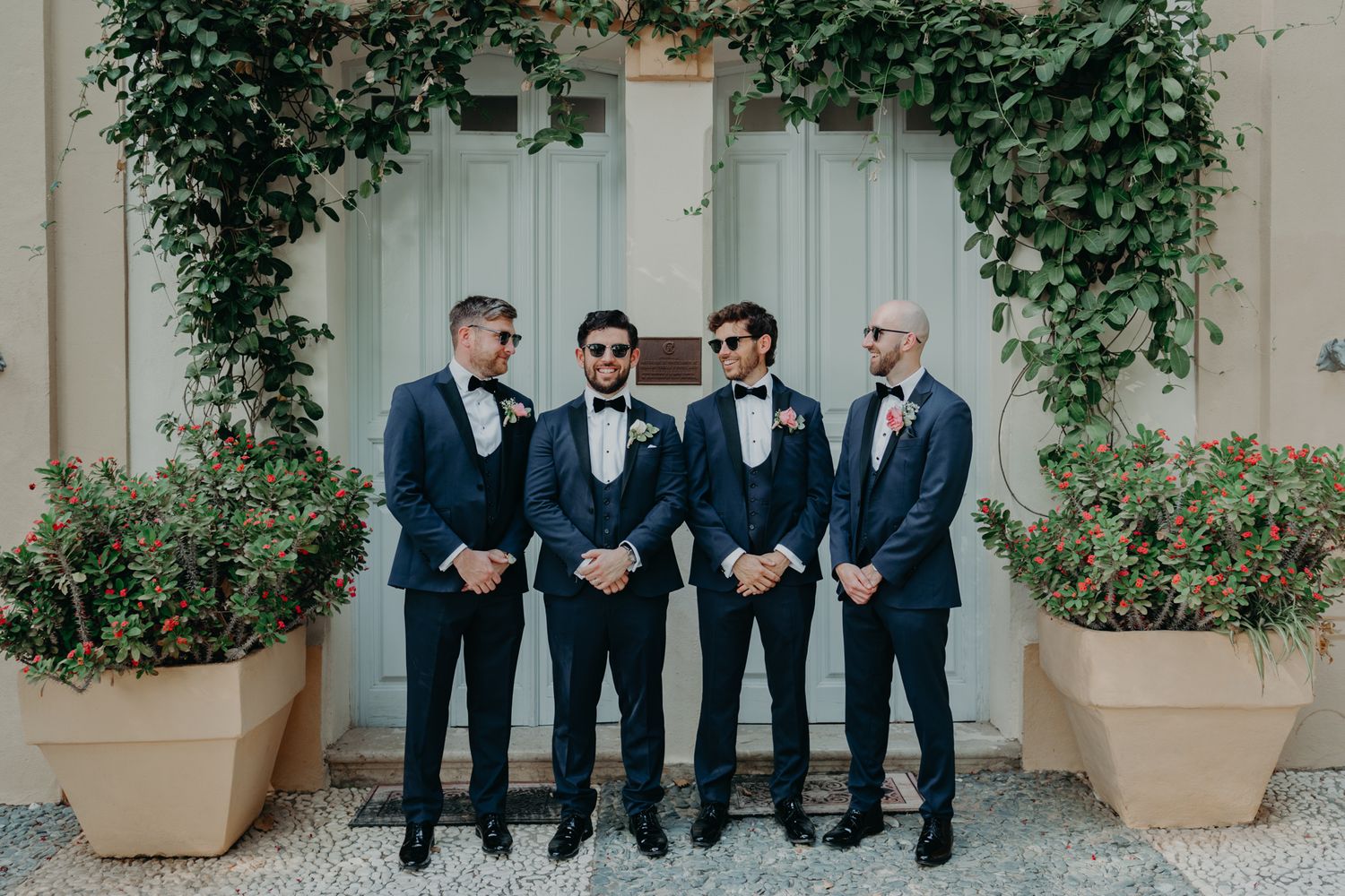 Four men in formal navy suits and bow ties pose in front of an ivy-covered wall with potted plants.