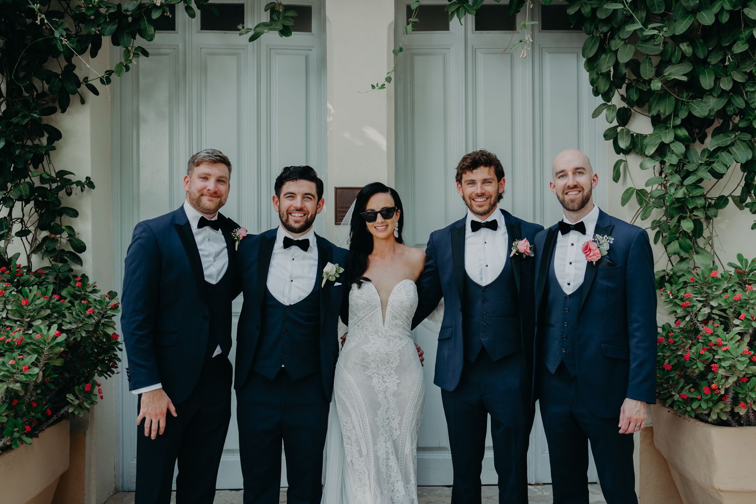 Wedding party in formal navy suits and gowns pose together against a light colored wall with climbing vines.