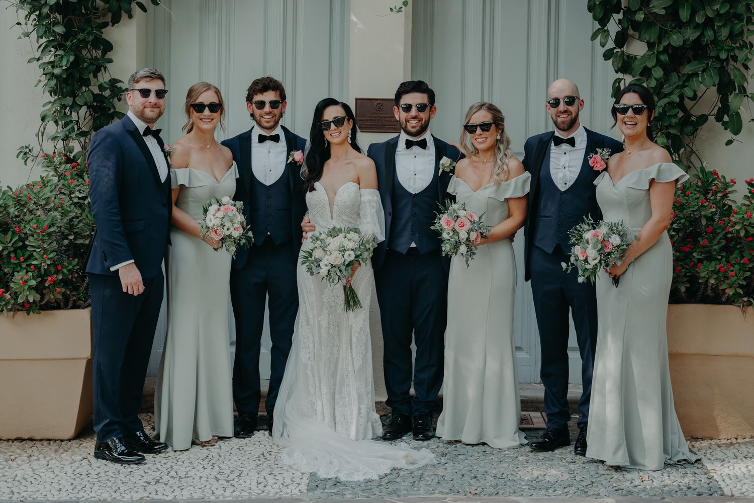 Wedding party in formal attire poses against white wall with greenery.