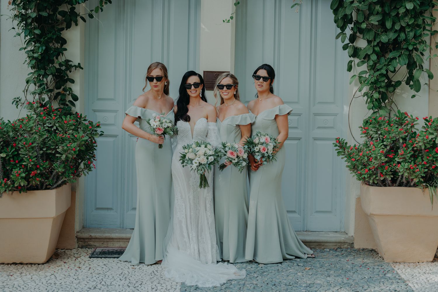 Four women in elegant mint bridesmaid dresses and sunglasses pose together against a light blue door with floral decor.