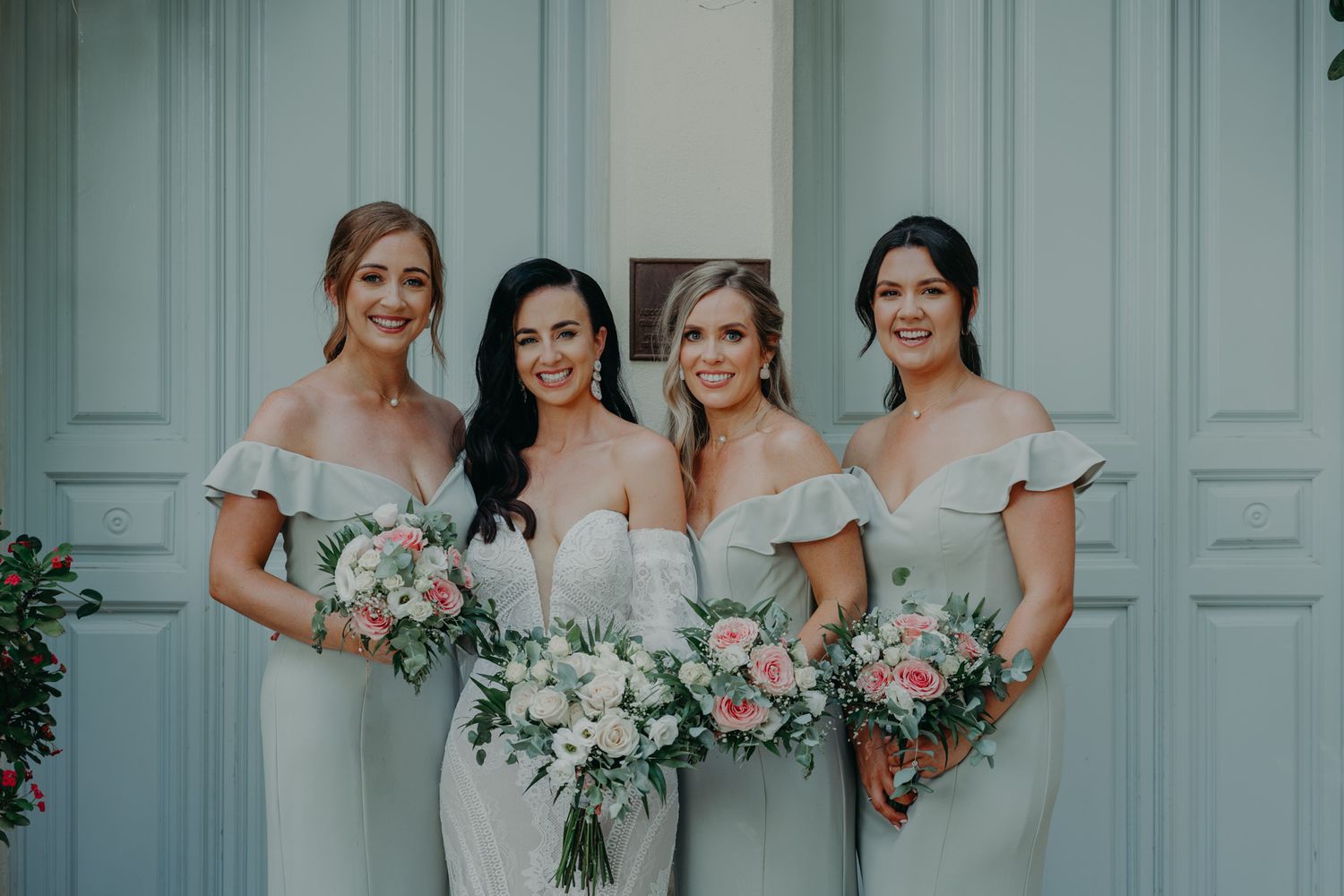 Four bridesmaids in elegant dresses holding beautiful pastel floral bouquets against a blue wall.