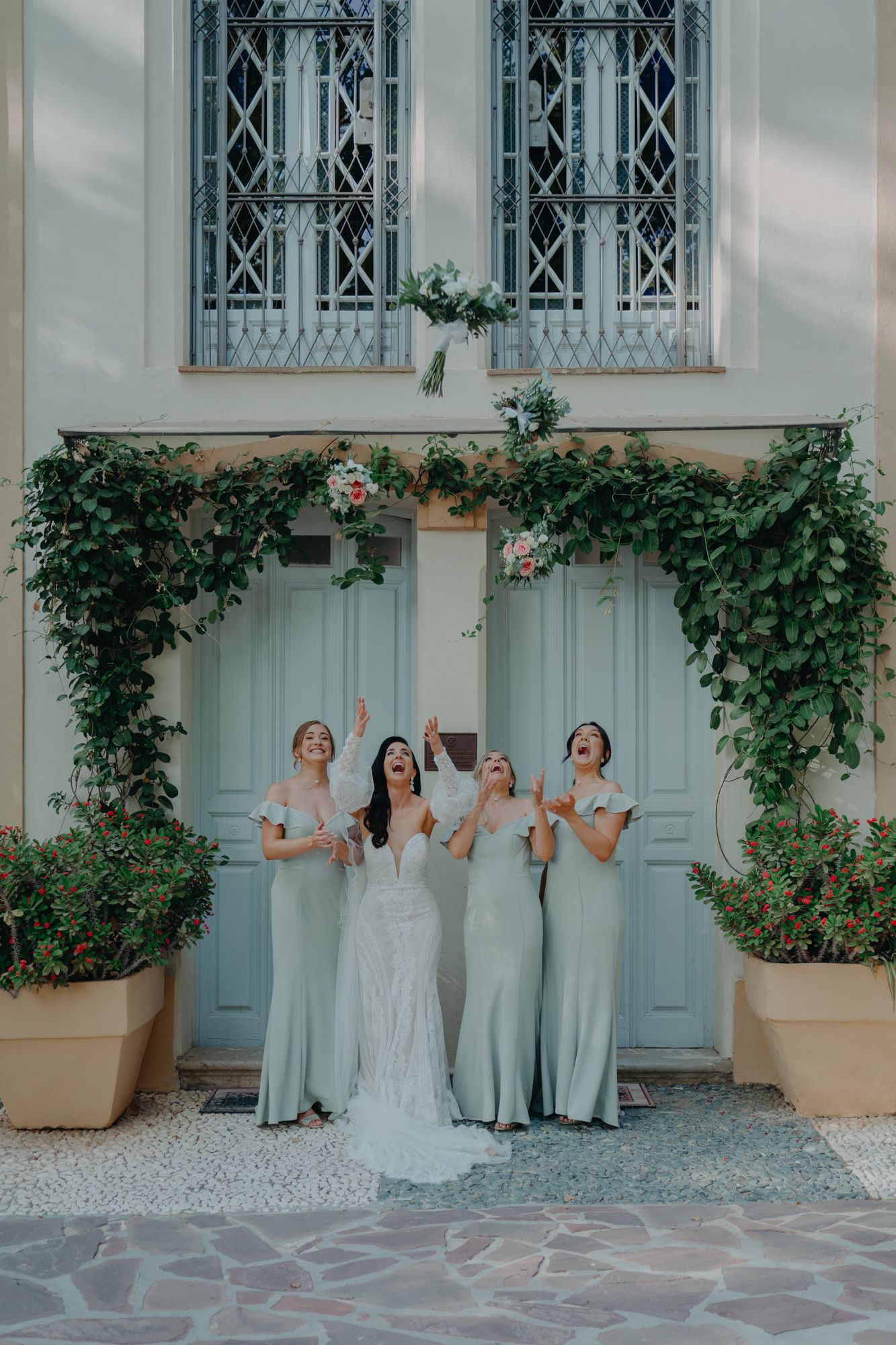A bridal party in sage green dresses poses in front of a light blue door with climbing vines and decorative windows.