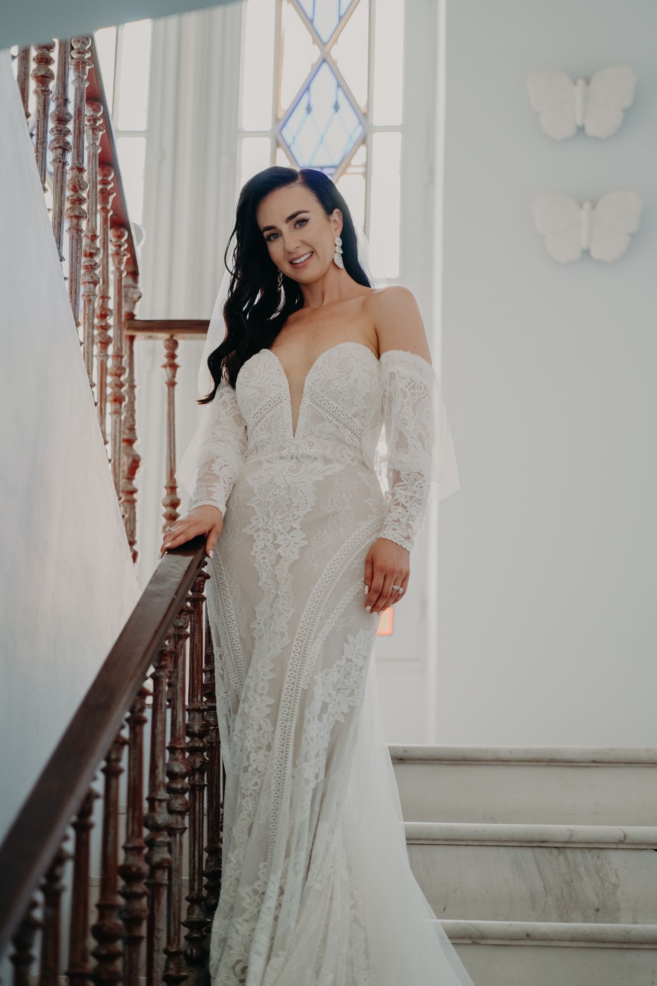 Stunning bride in a white lace off-shoulder wedding gown poses elegantly on vintage staircase with decorative railing.