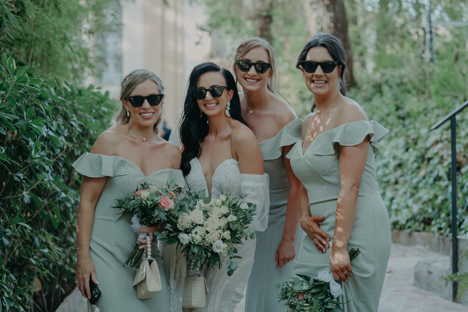 Wedding party in sage green dresses pose together while holding white and purple floral bouquets in a garden setting.