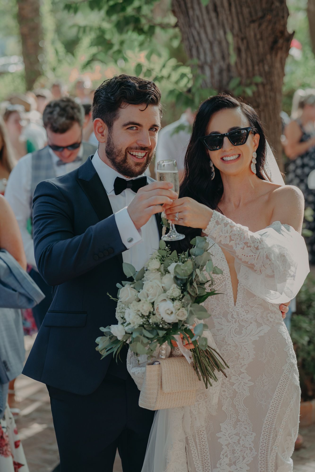A joyful wedding celebration outdoors with champagne toast and white floral bouquet.