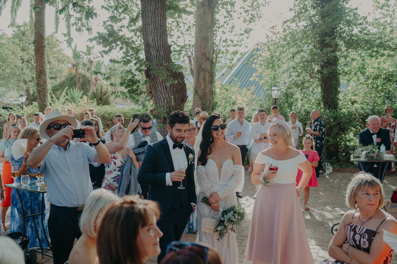 Wedding guests gather outdoors under trees to celebrate during an outdoor ceremony.