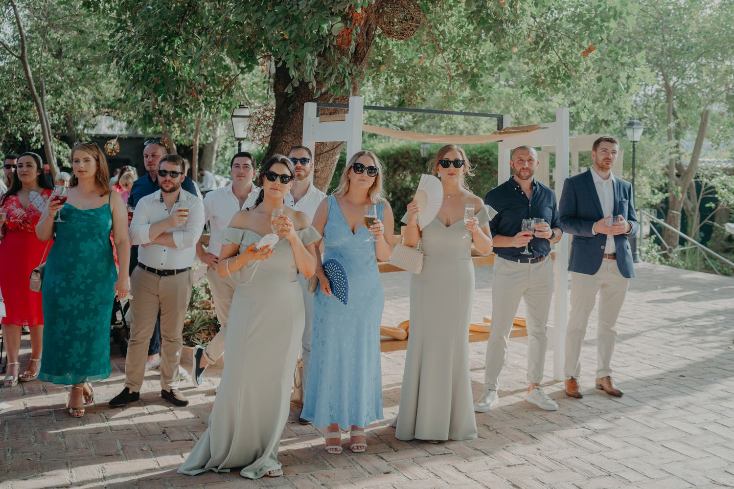 Wedding guests in summer attire stand in line wearing sunglasses at outdoor celebration.