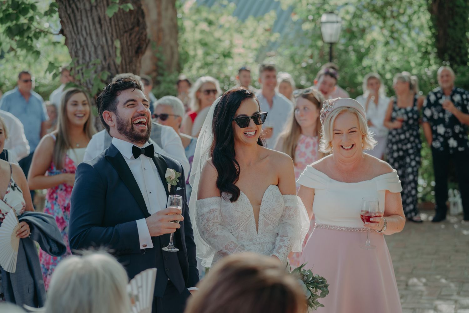 Wedding guests celebrating outdoors under trees during a sunlit ceremony.