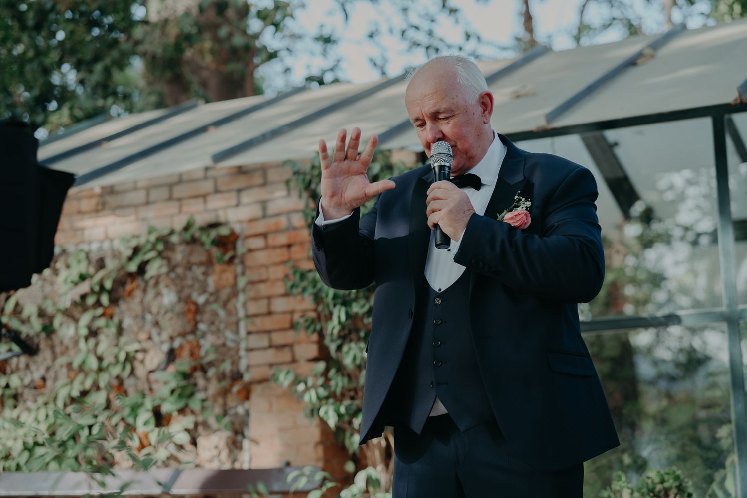 Speaker in formal attire delivers emotional speech during outdoor wedding reception.