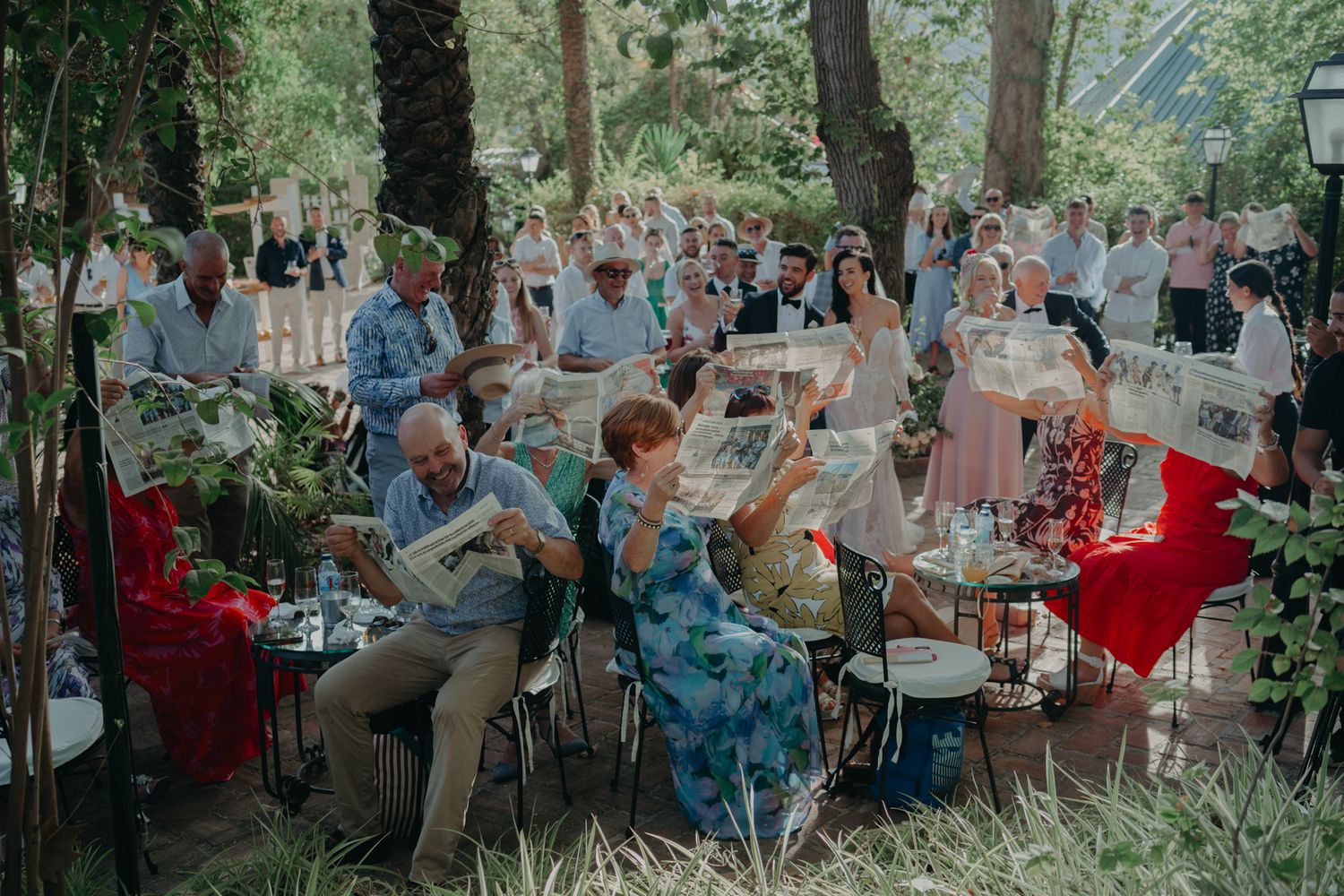 Wedding guests seated in an outdoor garden setting raise their glasses in celebration.