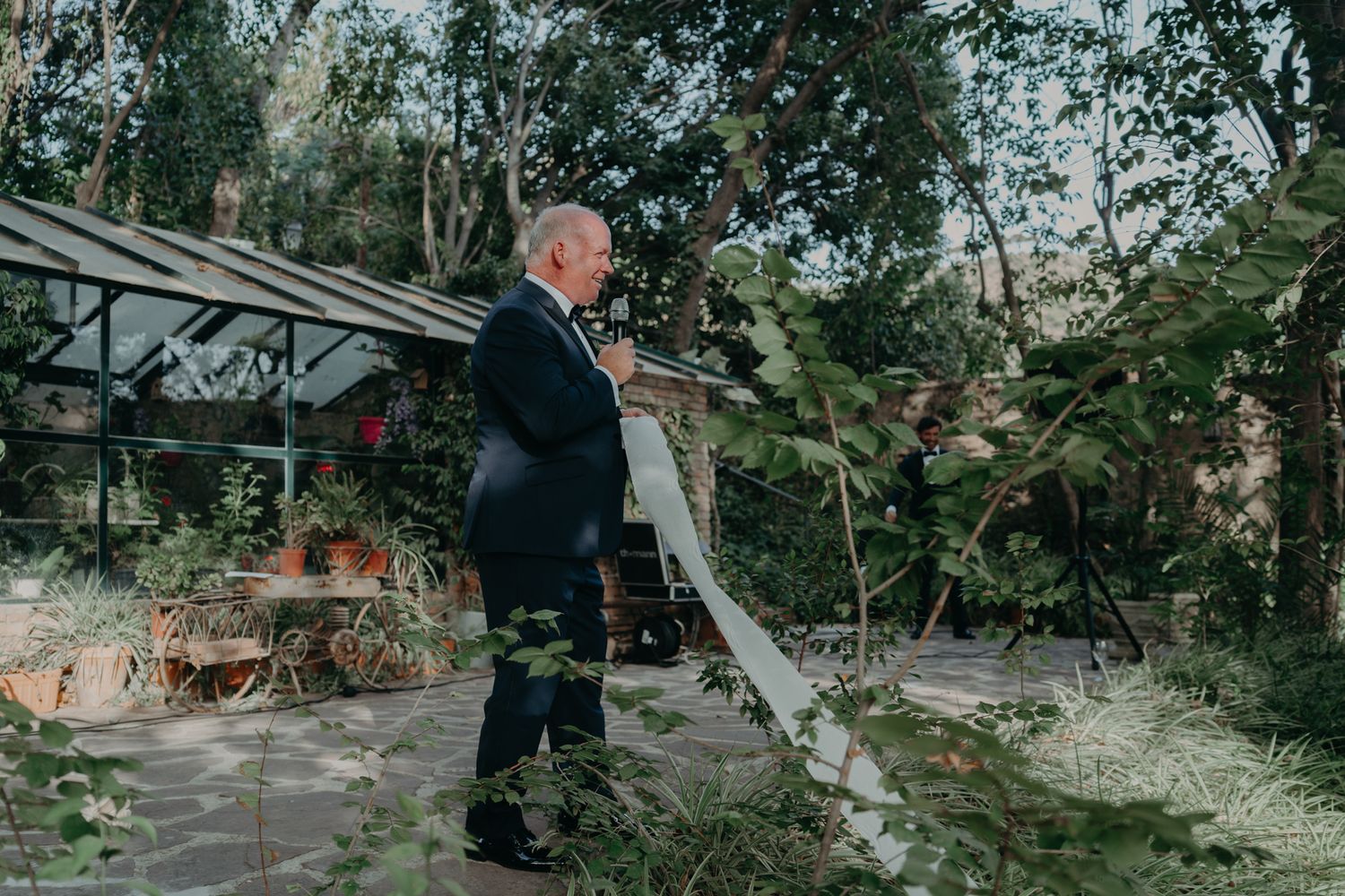 Person in formal attire stands thoughtfully in garden setting with brick wall and trees.
