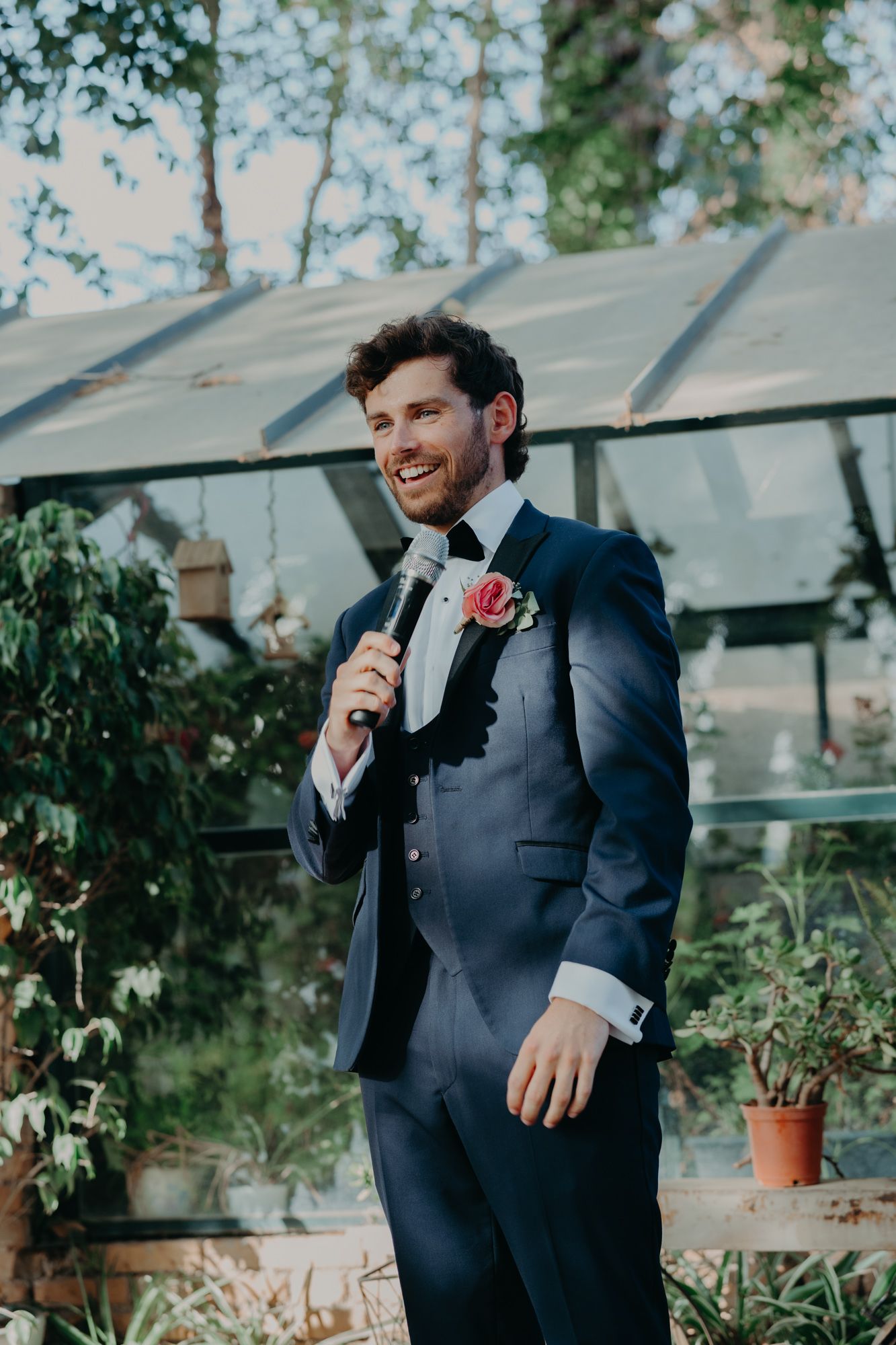 Formal portrait of suited individual with pink boutonniere in front of greenhouse.