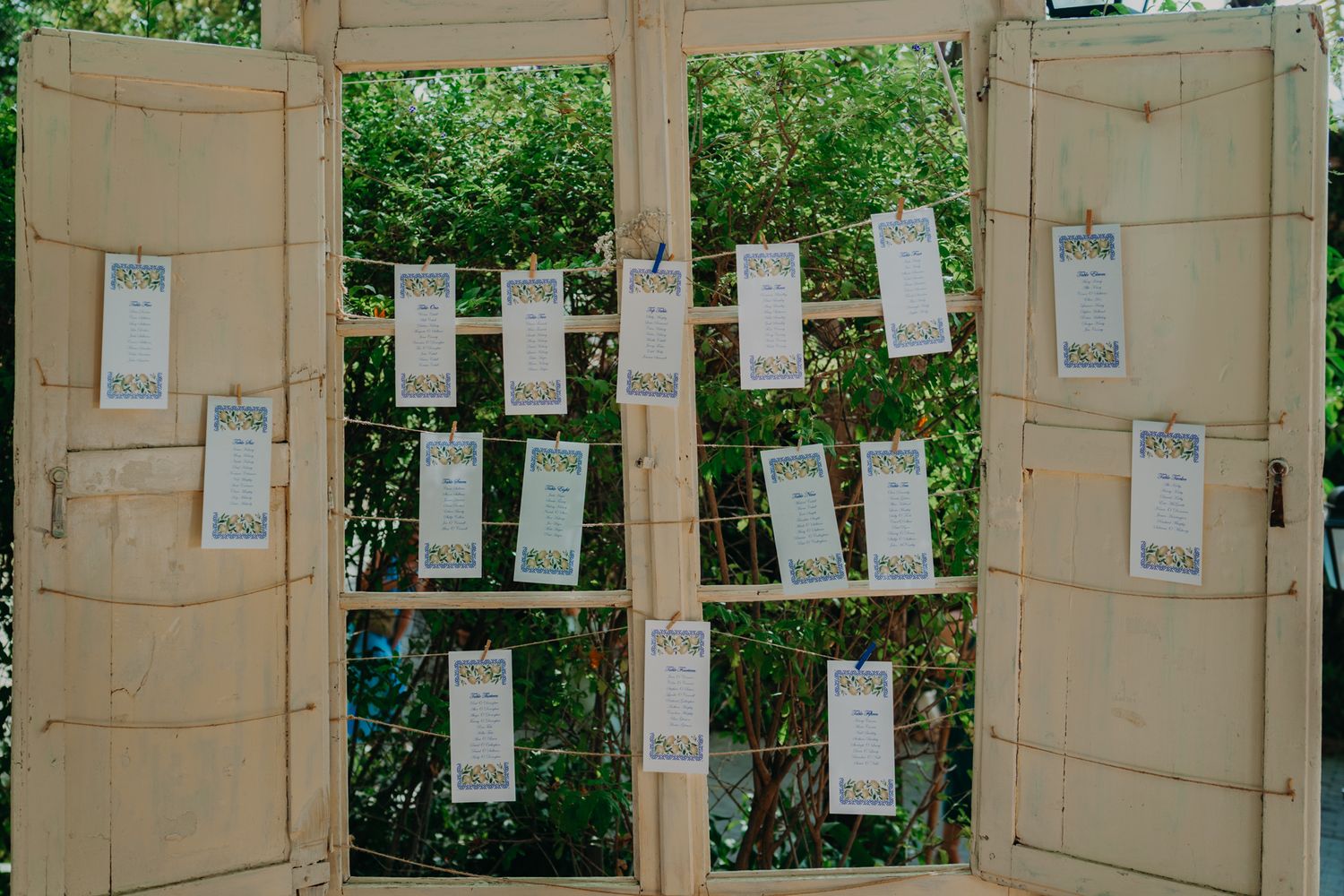 White paper seating charts hang from strings between two vintage wooden doors against a green garden backdrop.