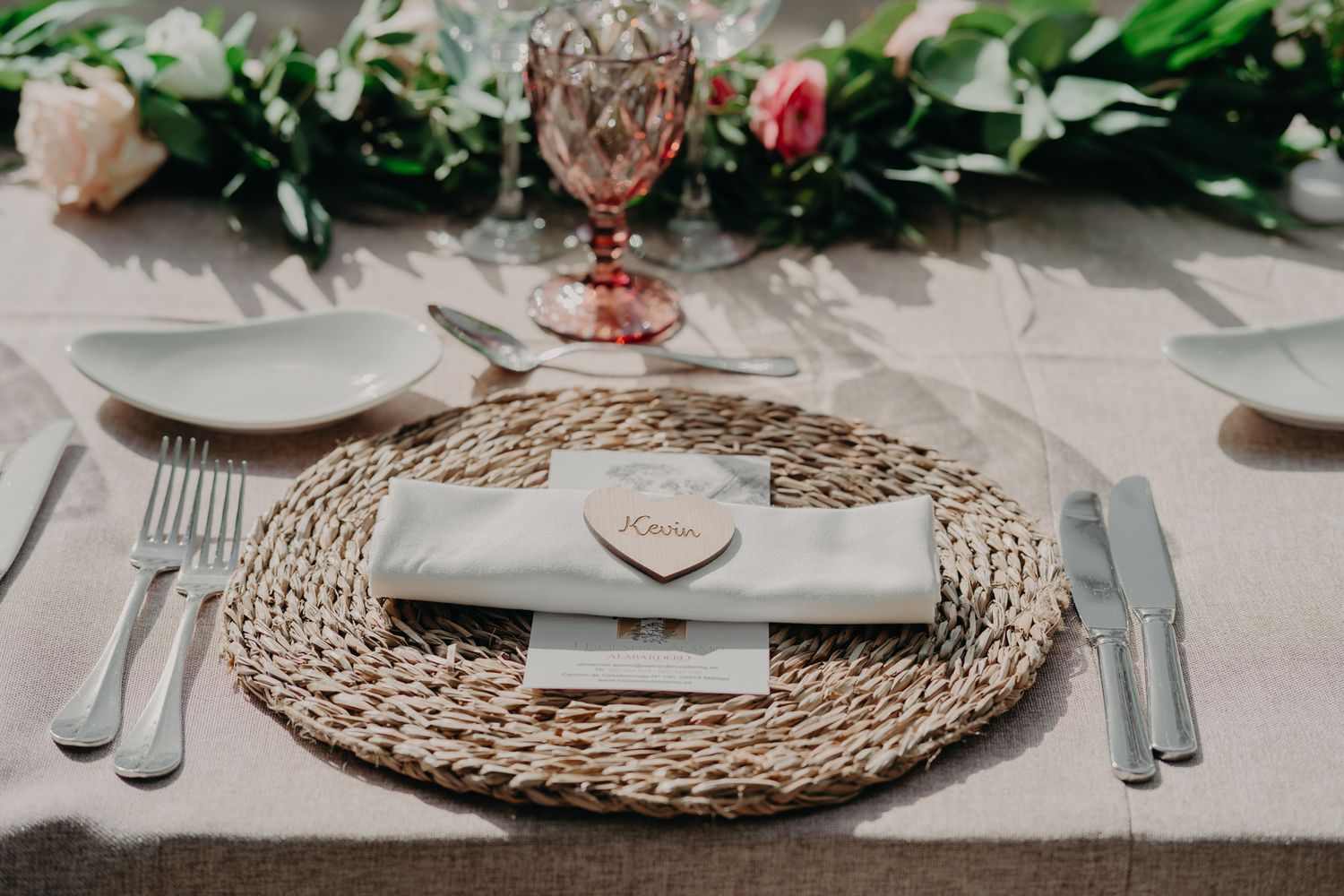 An elegant place setting with woven charger plate, cutlery, and wine glass on a neutral tablecloth.