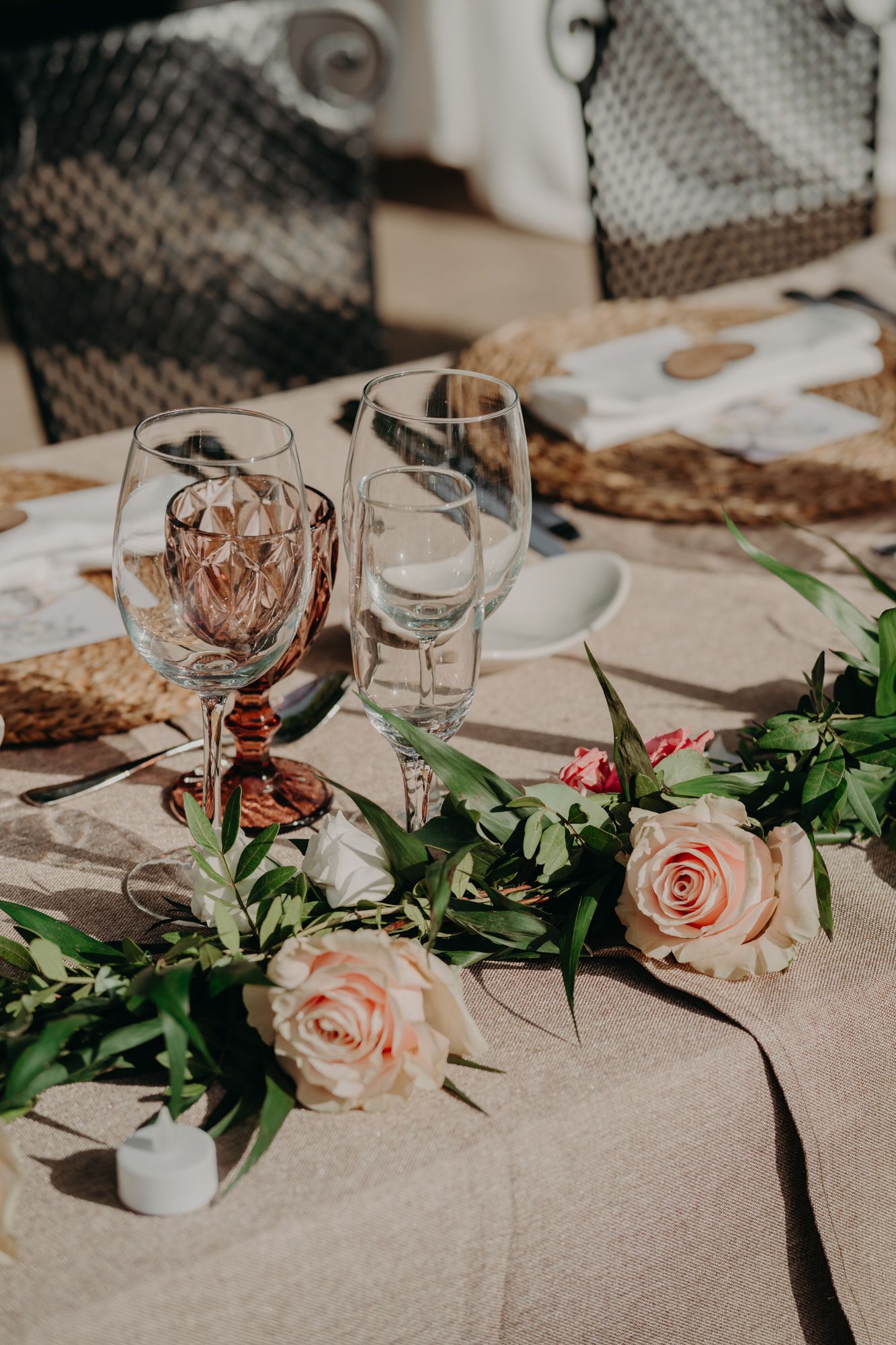 Elegant table setting with wine glasses, pink roses and greenery on a beige tablecloth.