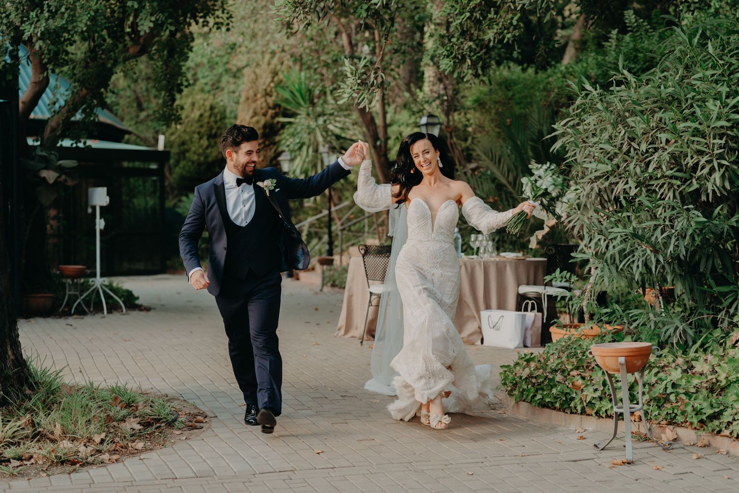 A couple in wedding attire walks along a garden path at an outdoor venue during their celebration.