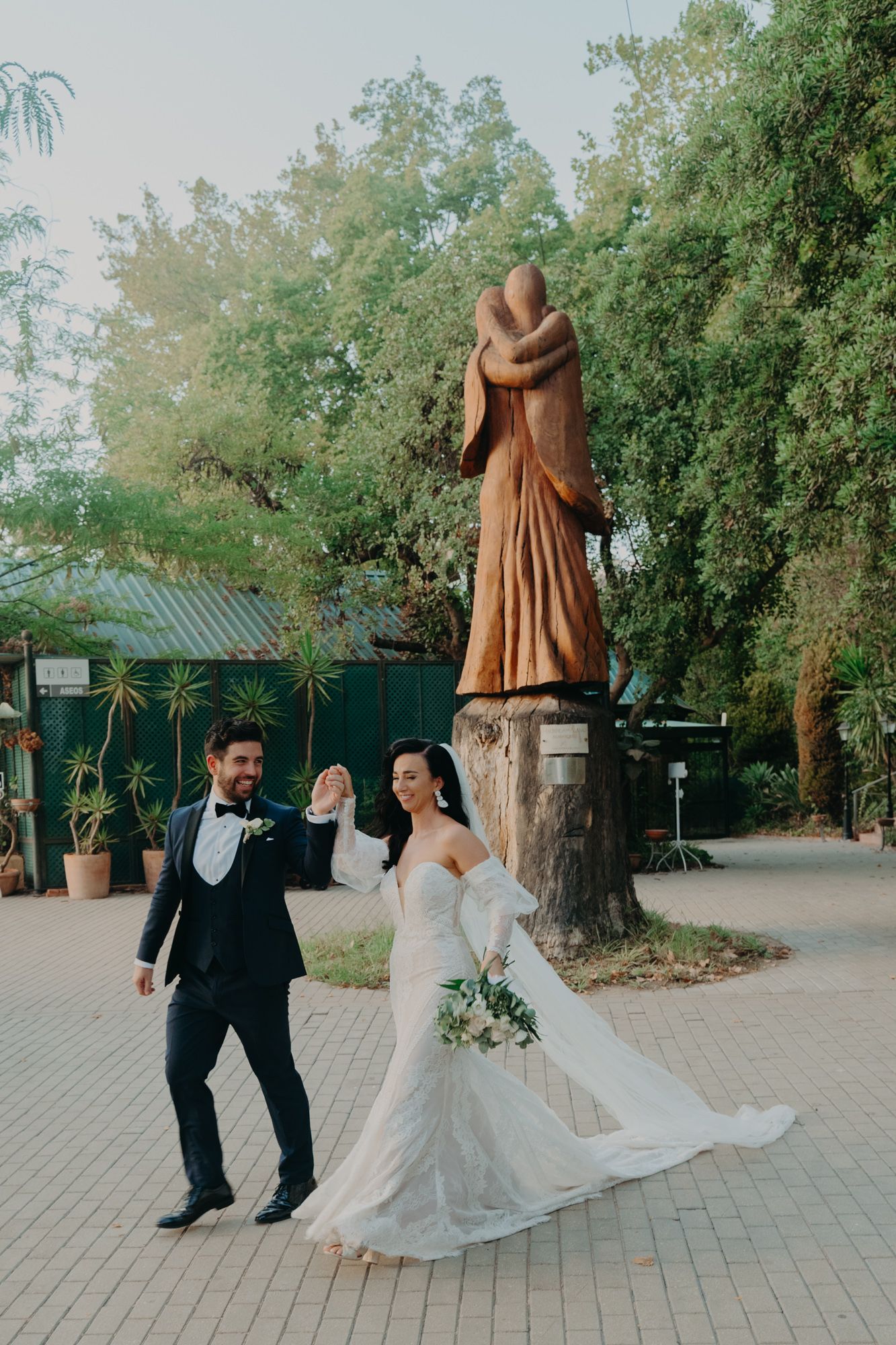 Wedding couple celebrating near a large statue in a garden courtyard setting with lush trees and greenery.