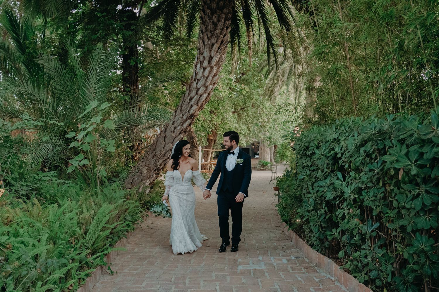 Newlyweds walking hand in hand along a palm-lined garden path.