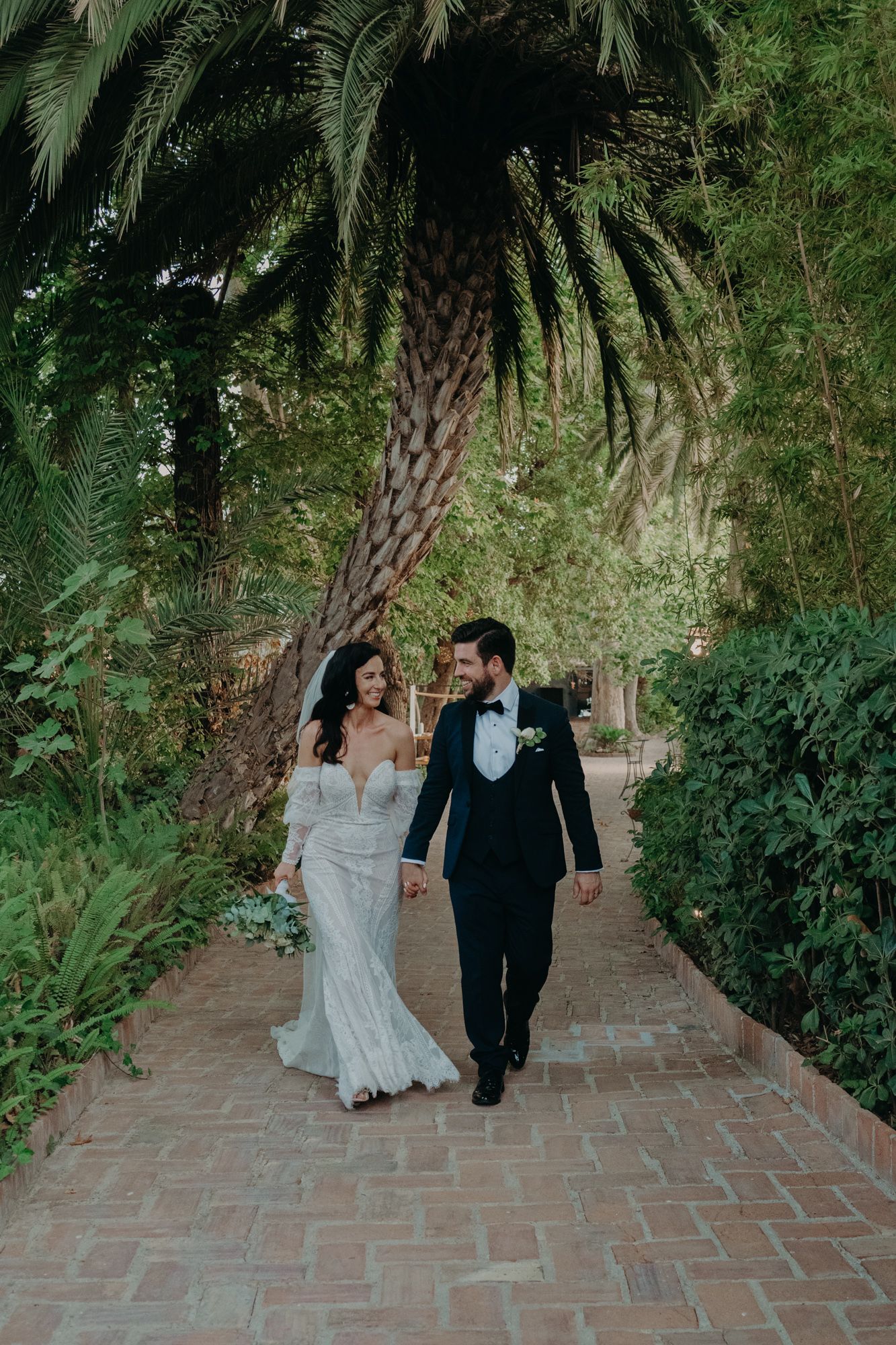 A wedding couple walks down a palm tree-lined brick pathway in formal attire.