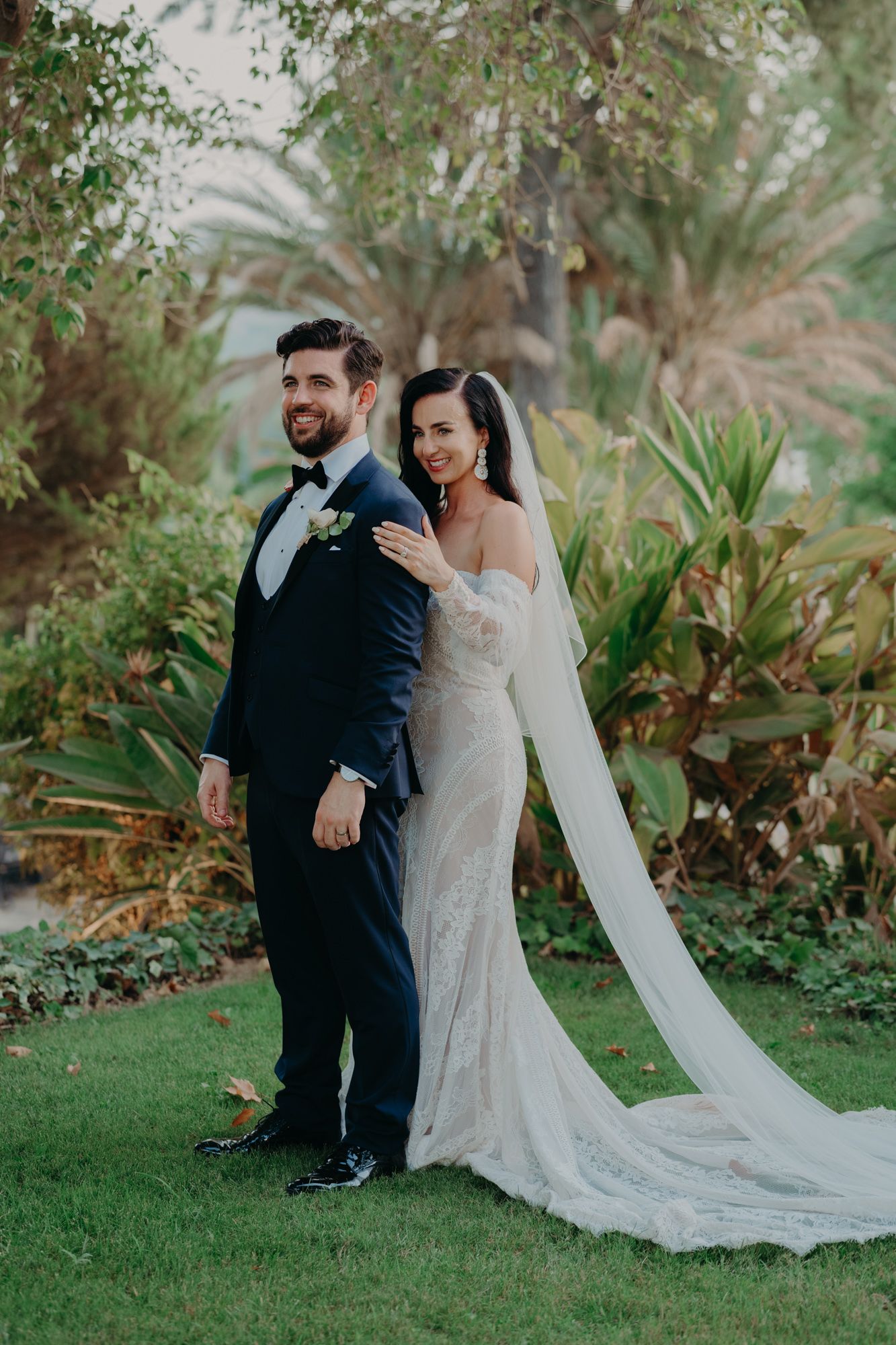 A wedding couple poses together on a lawn with palm trees and tropical foliage in the background.
