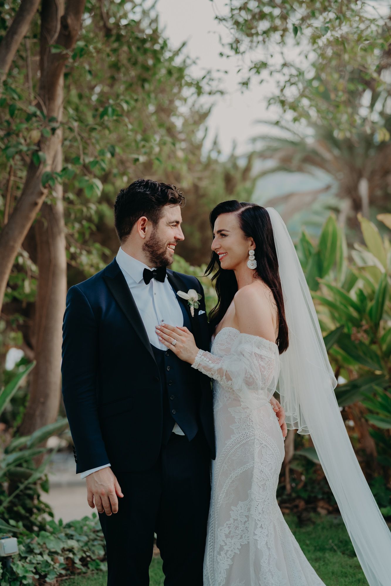 Romantic wedding portrait in lush garden setting with flowing veil and formal attire.