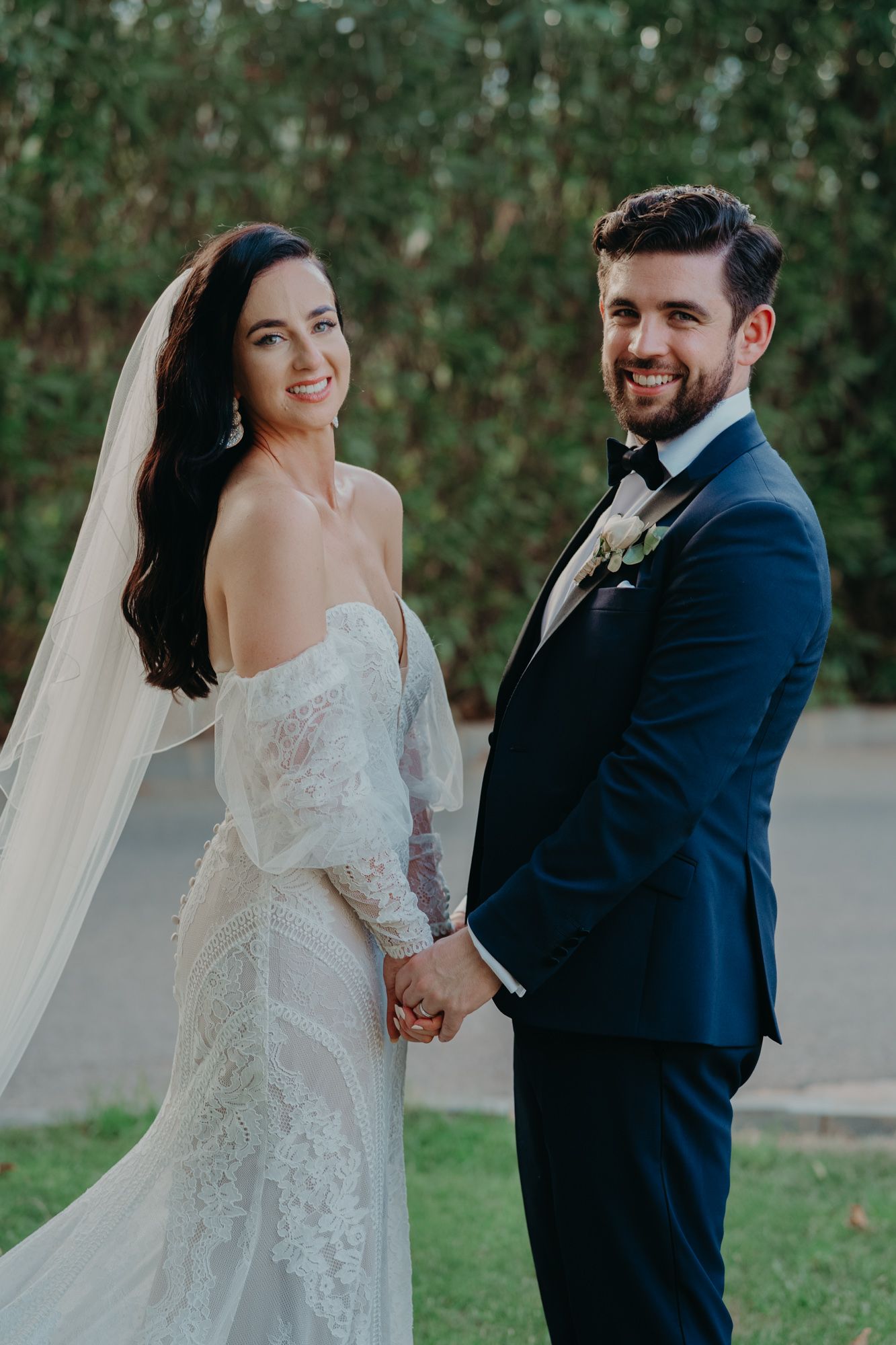 Happy newlyweds share a romantic moment in a garden setting, bride in lace gown and groom in navy suit.