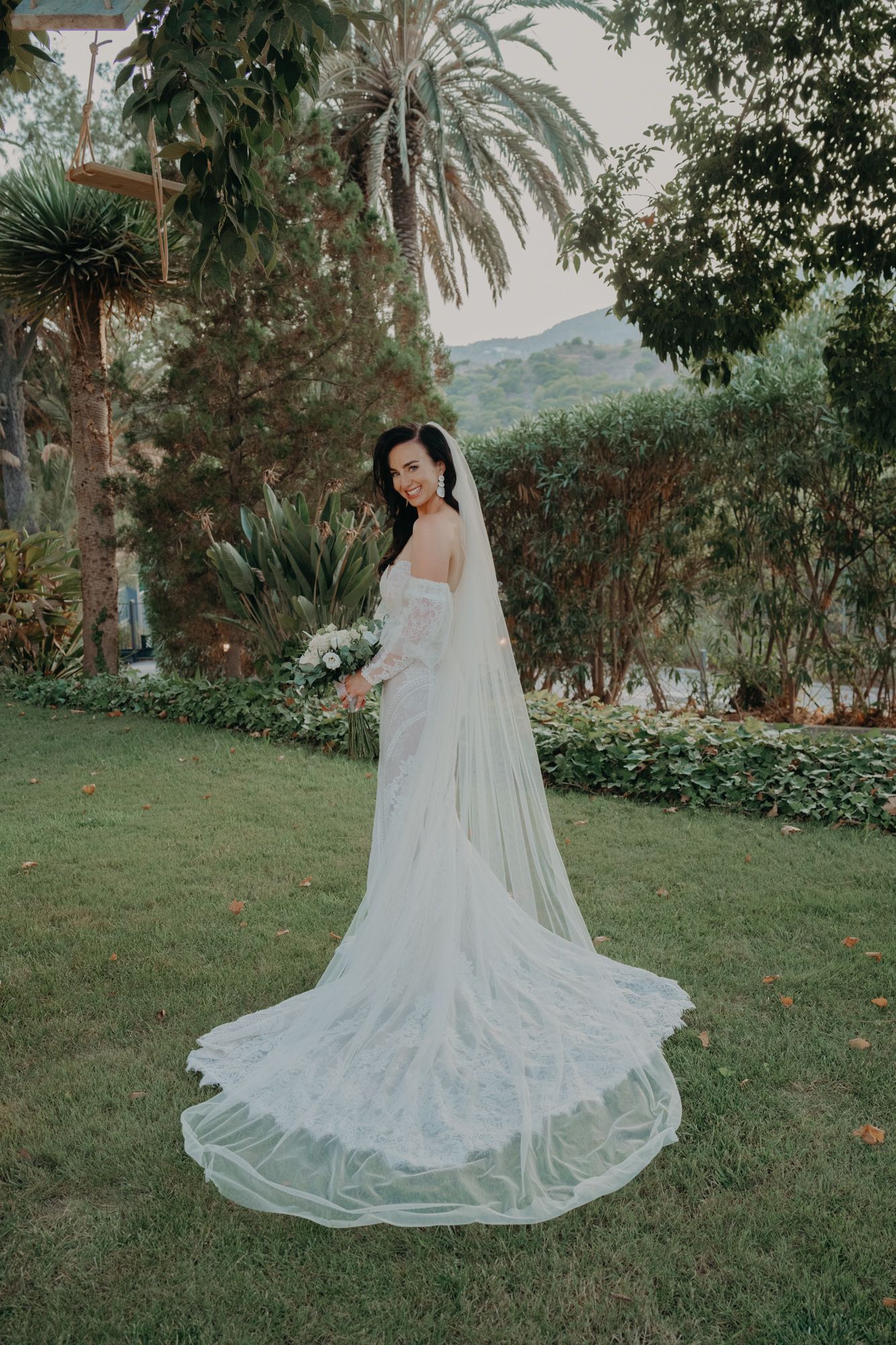 Bride in elegant white lace wedding gown with long flowing veil poses in garden setting with palm trees and mountains.