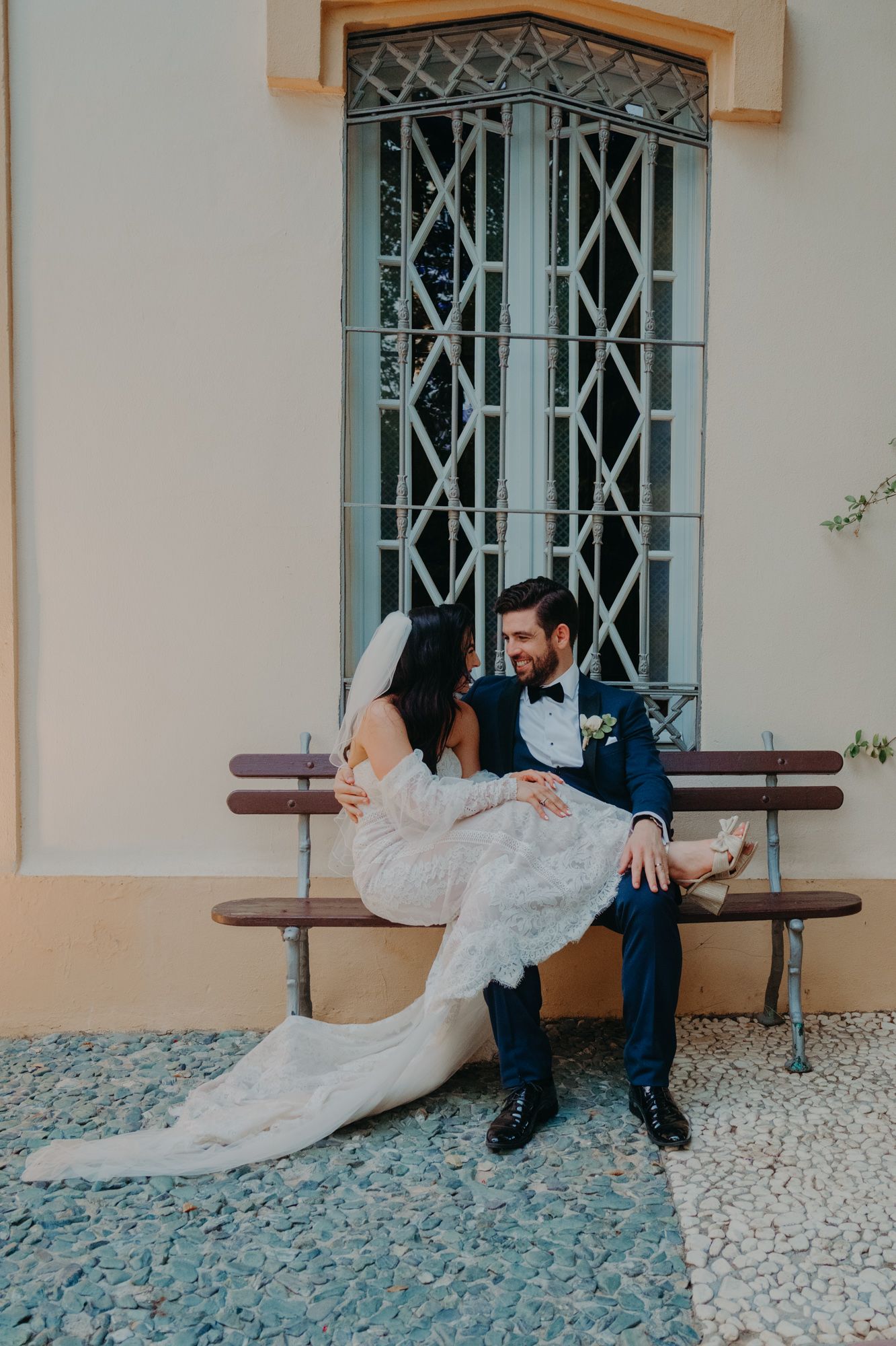 Newlyweds share romantic moment on bench by ornate window with bride's dress gracefully draped.