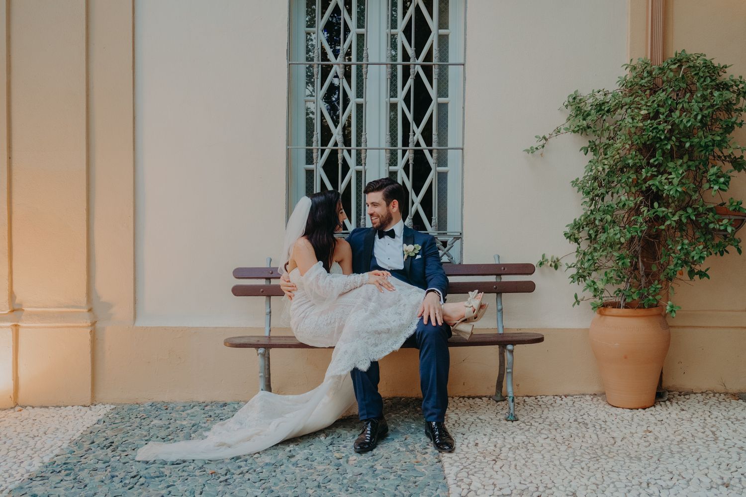 A couple shares an intimate moment on a wooden bench outside a building with ornate windows and potted plants.