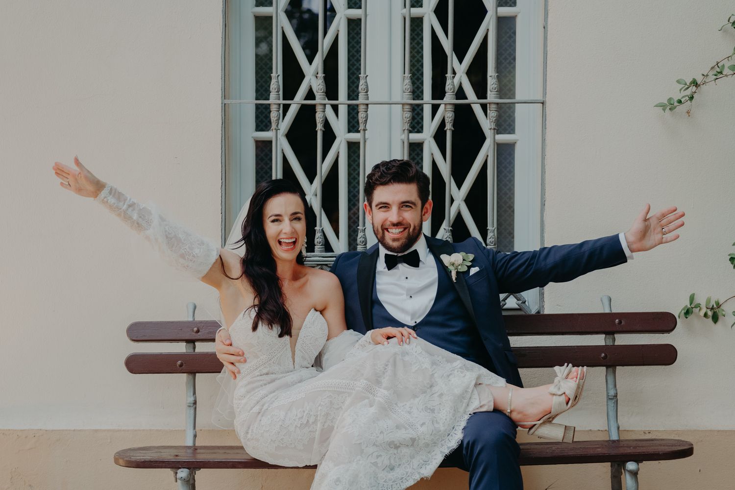 Newlyweds sitting playfully on a bench with arms outstretched against a white building.