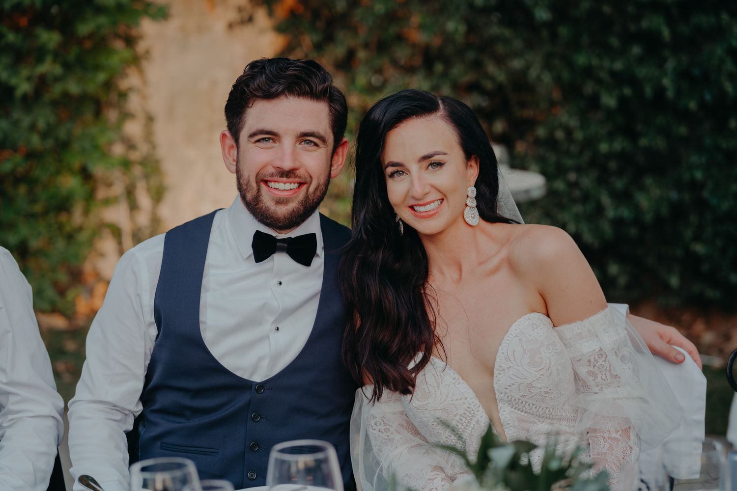 A newlywed couple smiles while seated at an outdoor reception table.