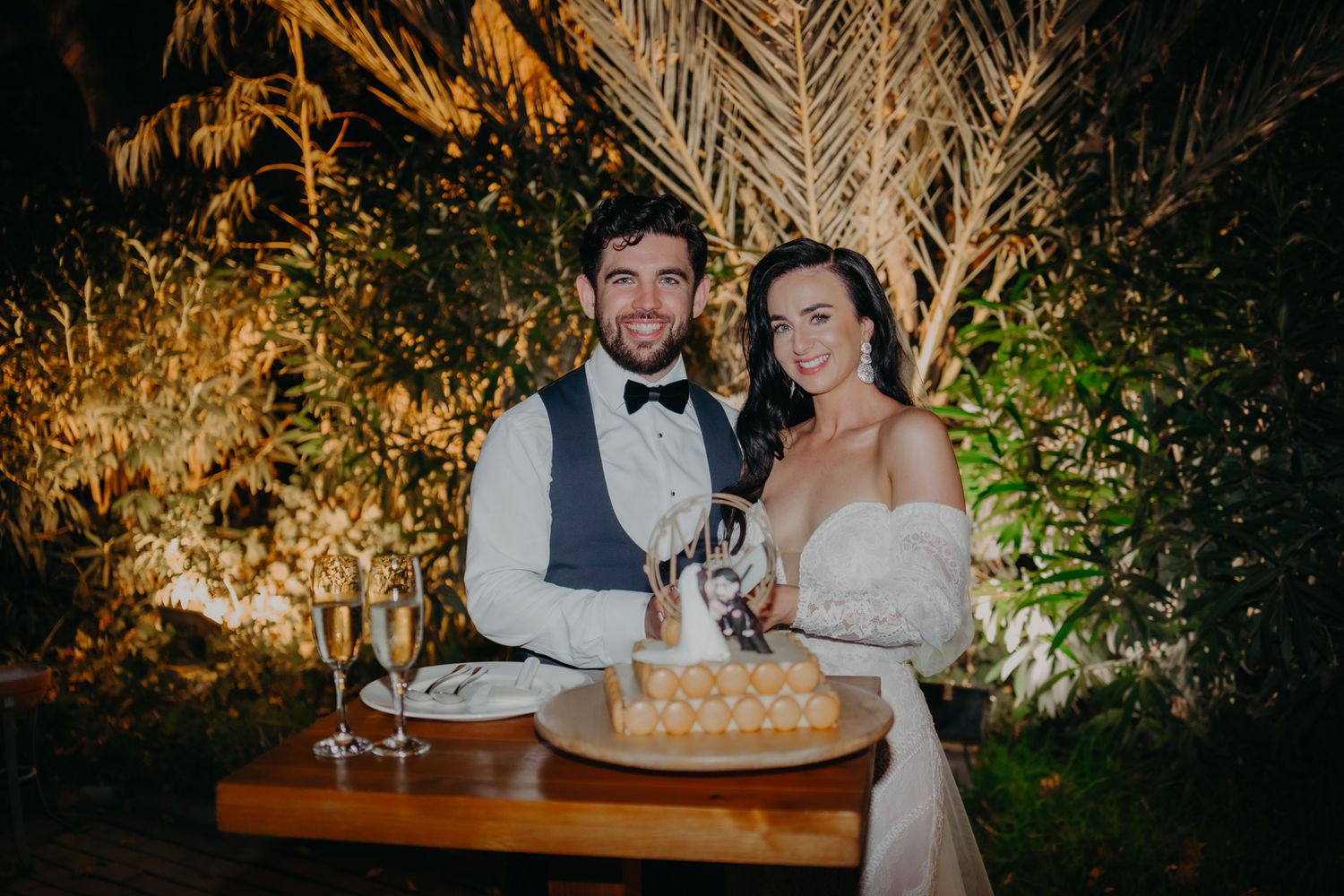 Newlywed couple cuts their elegant white wedding cake together at an outdoor evening reception with palm trees.