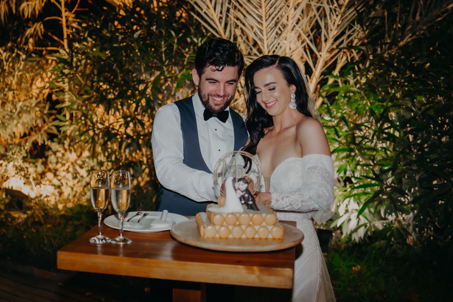 A wedding couple cuts their cake together at an outdoor evening reception with illuminated foliage in the background.