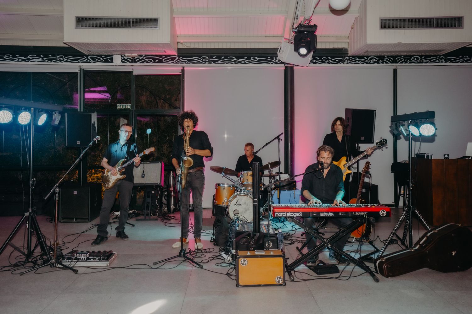 Musicians perform on stage with instruments and equipment in a modern studio space with colorful lighting.