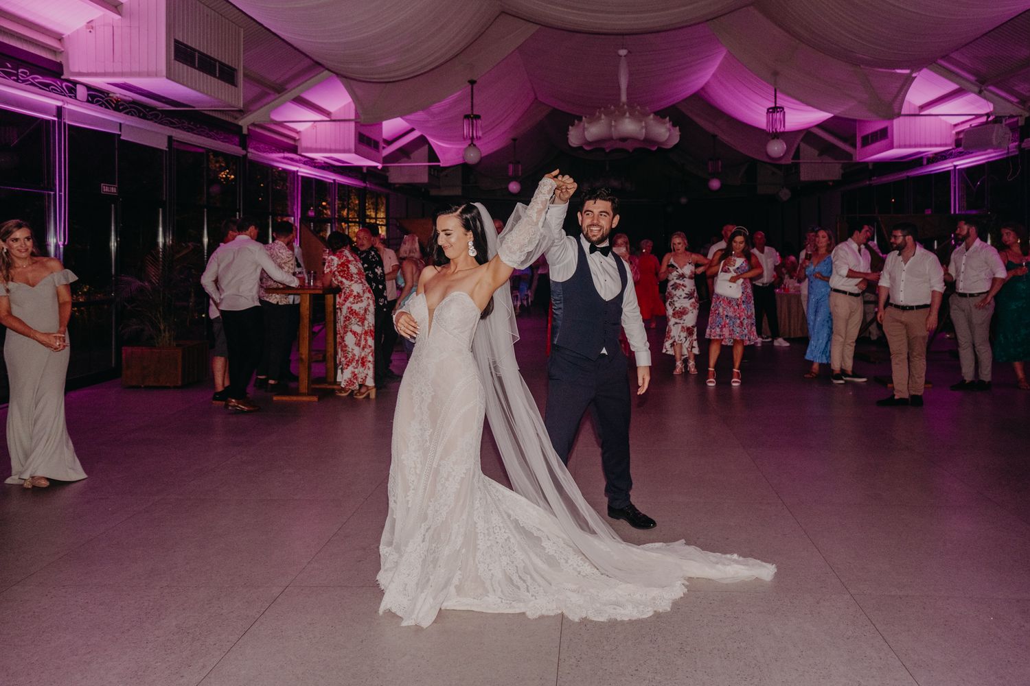 Couple dances in a ballroom under magenta lighting while guests watch from the sidelines during a wedding reception.