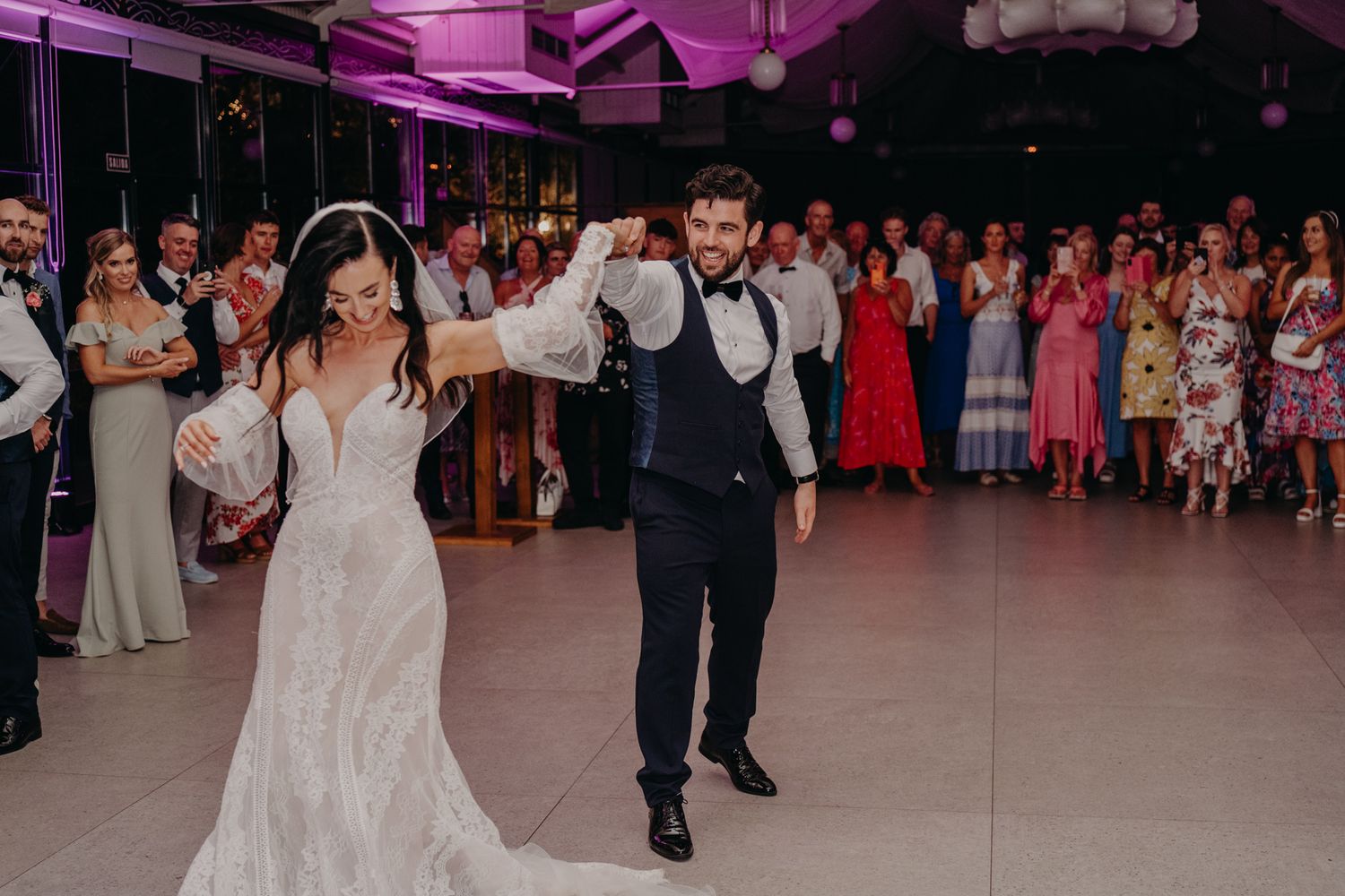 A couple performs their first dance at their wedding reception under dramatic pink lighting while guests watch.