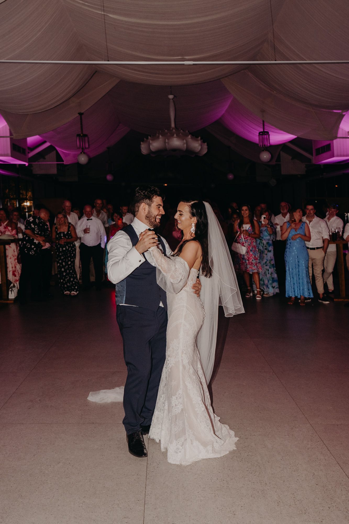 Newlyweds share their first dance at a wedding reception under purple mood lighting and draped fabric ceiling.