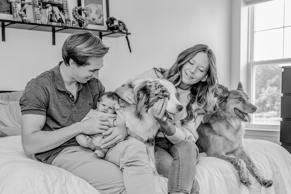 Black and white photo of a couple sitting on a couch snuggling with their two dogs in a bright room.