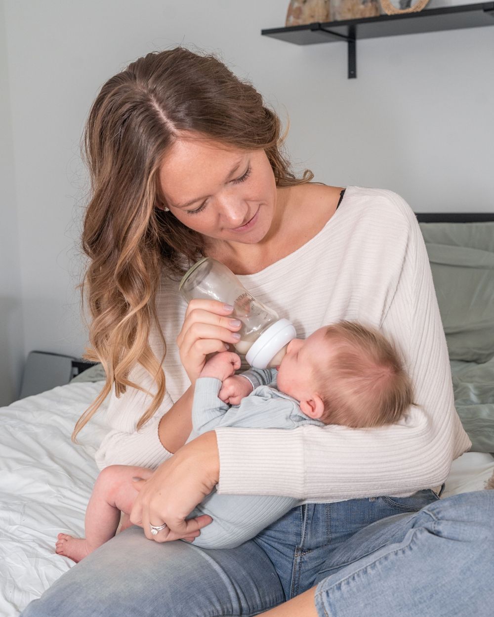 A parent bottle feeds a baby while sitting comfortably on a bed wearing a cream sweater and jeans.