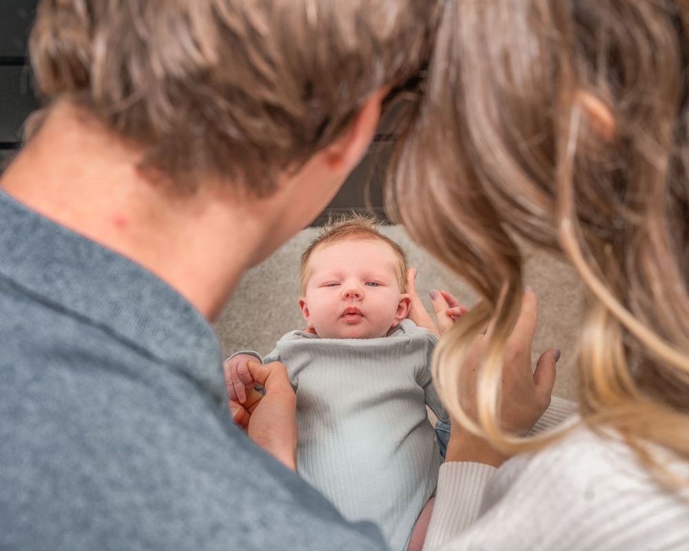 A newborn baby wrapped in a grey blanket is held between two people wearing grey and beige clothing.