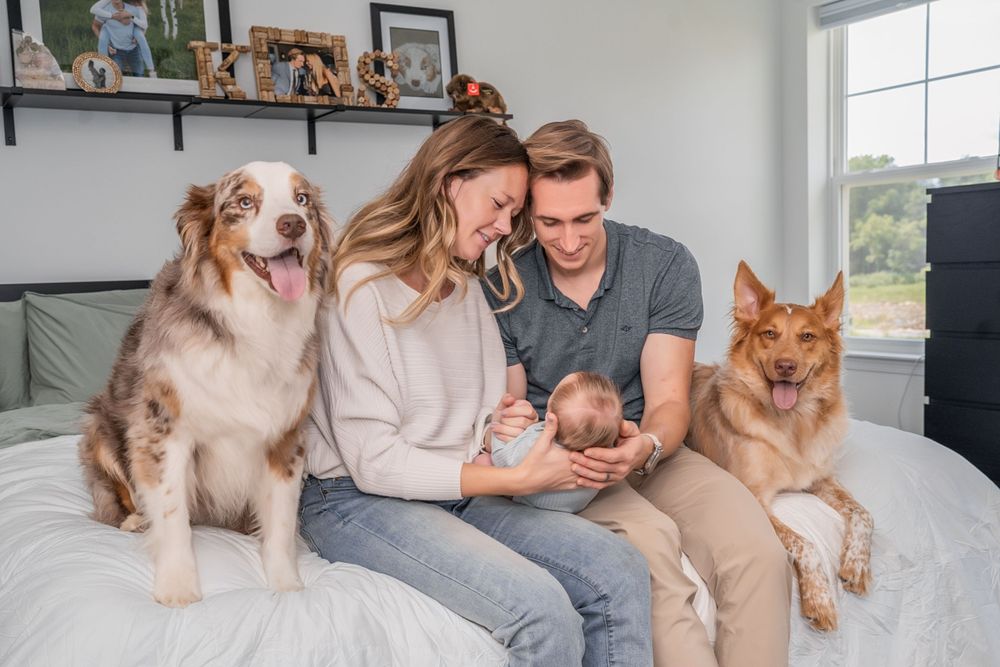 Family and two dogs sitting together on couch with newborn baby in a bright living room.