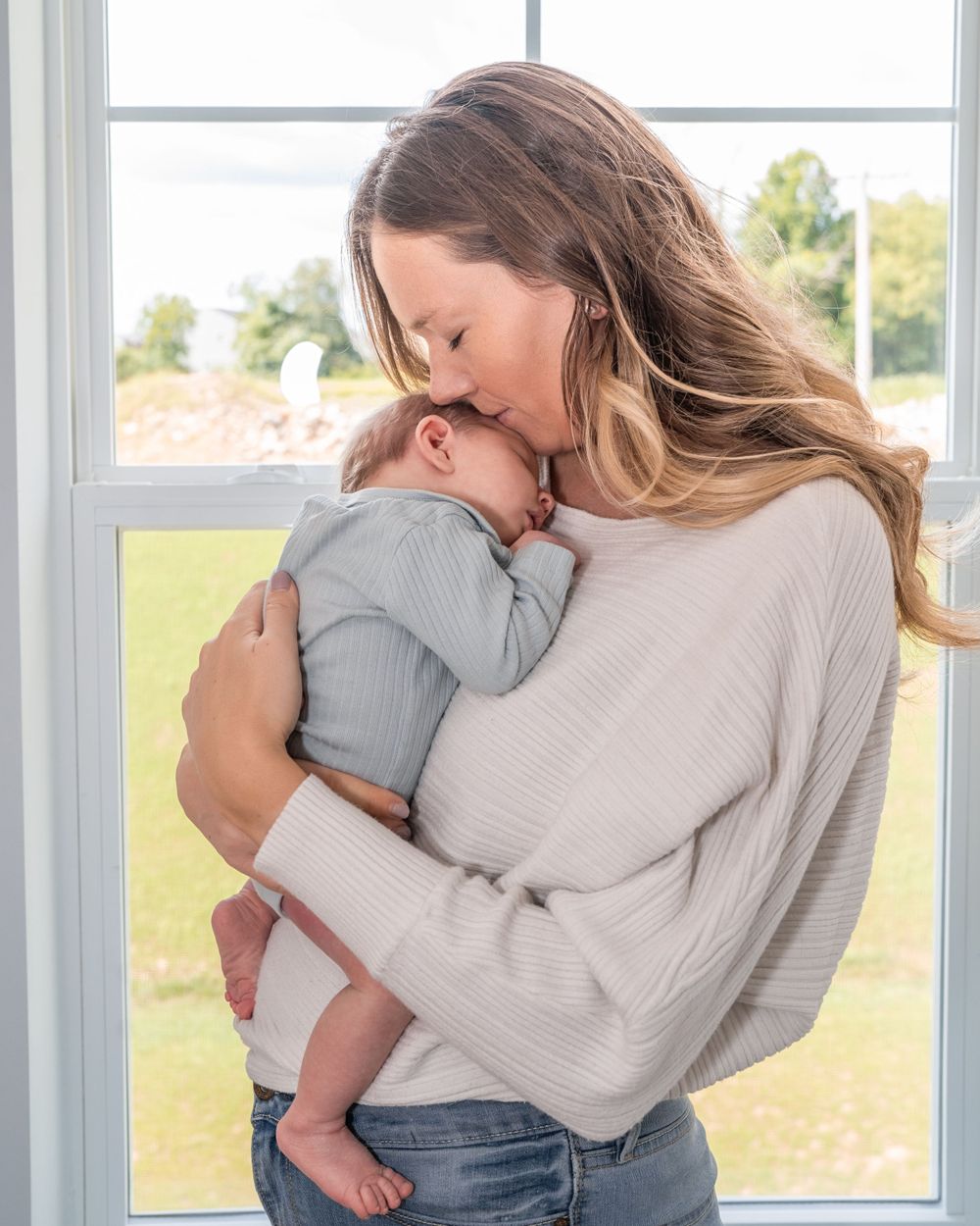 A parent in a cream sweater holds and embraces their infant tenderly by a sunny window.