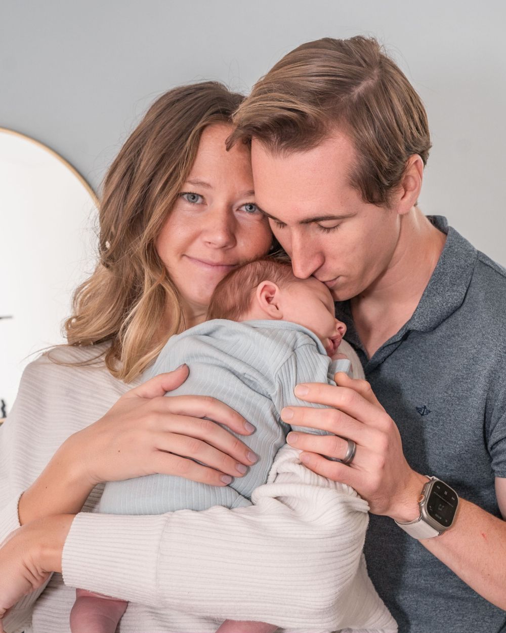 A tender family moment as parents embrace their newborn baby wrapped in a light grey outfit.