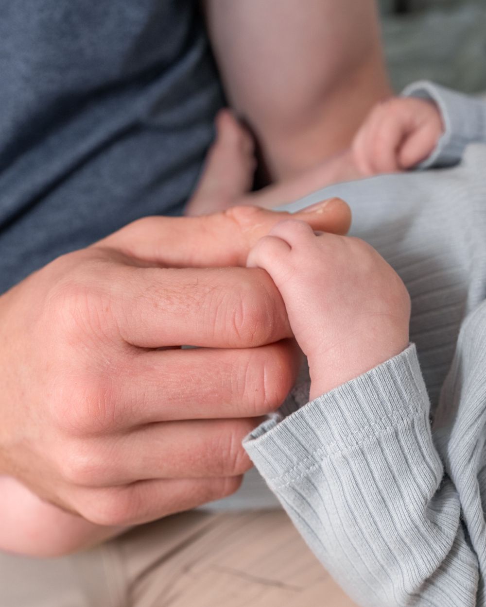 Close-up of hands gently holding a tiny baby's hand in a grey sweater sleeve.